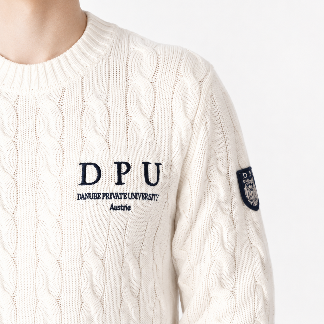 DPU Strickpullover in Winterweiß mit klassischem Zopfmuster, blauem DPU-Stick und aufgenähtem Wappen-Patch am linken Ärmel.