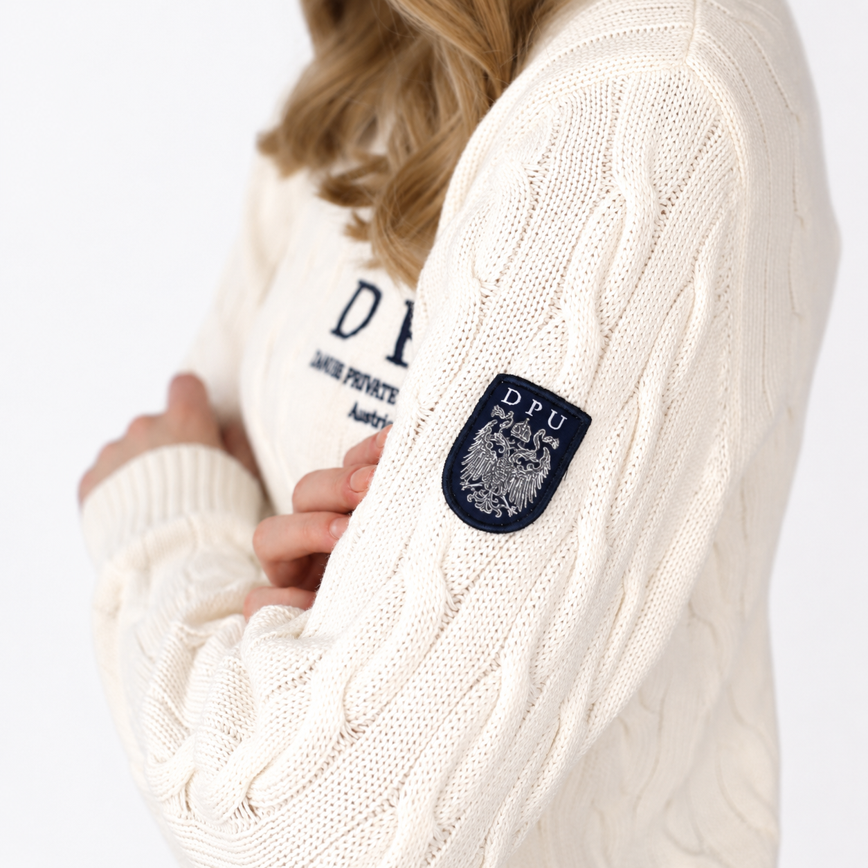 DPU Strickpullover in Winterweiß mit klassischem Zopfmuster, blauem DPU-Stick und aufgenähtem Wappen-Patch am linken Ärmel.
