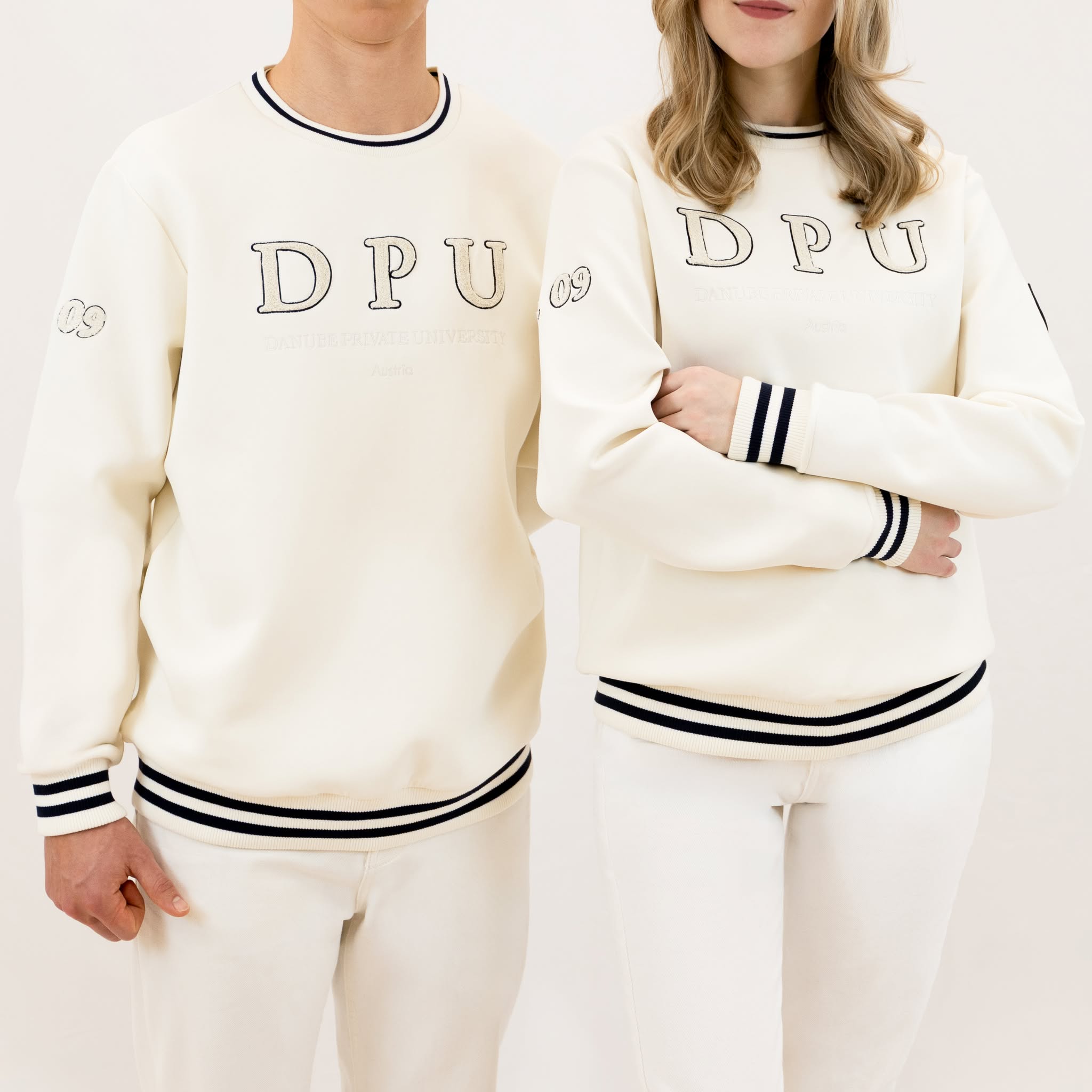 Winterweißes DPU-Sweatshirt mit Ton-in-Ton gesticktem Logo auf der Vorderseite, est. 09 am rechten Ärmel, Wappen-Patch am linken Ärmel und blauen Streifen an den Bündchen.